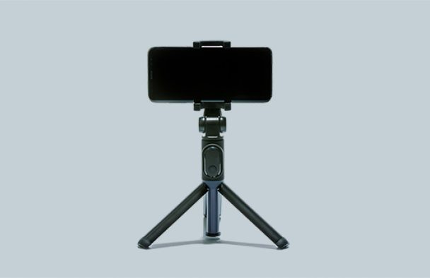 A smartphone tripod with a phone sits on a table with a grey background.