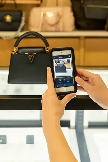 Person using a smartphone with the Entrupy app to authenticate a black Louis Vuitton handbag displayed on a glass counter in a luxury retail store.