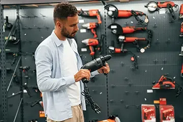 Man examining a power drill in a pawn shop with various red and black power tools displayed on a wall behind him.