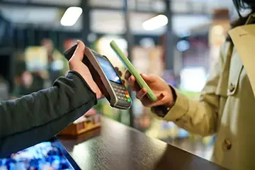 Customer making a contactless payment using a smartphone at a pawn shop, holding the phone near a card reader held by a cashier.