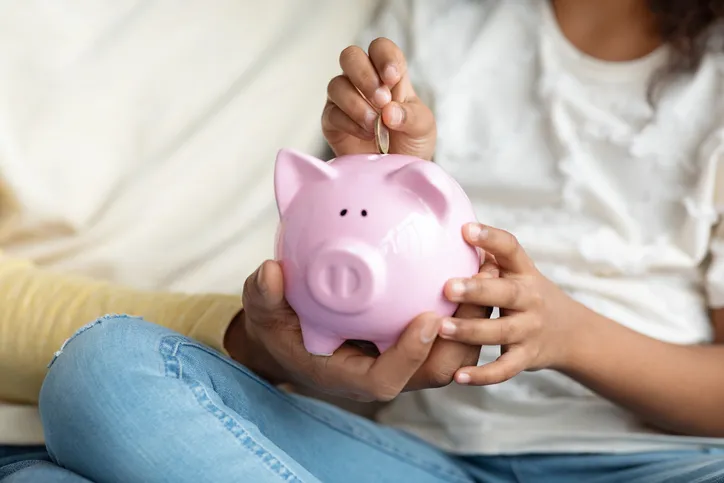 A close up picture of a black father and daughter sitting down while the daughter places a quarter in a pink piggybank.