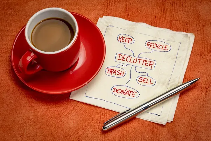 A cup of coffee sits on a saucer which sits on a napkin with handwriting on the napkin. The words “declutter,” “keep,” “recycle,” “trash,” “sell,” and “donate” are all handwritten on the napkin.