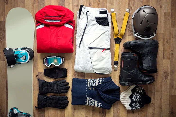 A bird’s eye view of snow gear someone would use while snowboarding: snowboard, goggles, gloves, hat, boots, gloves, helmet, suspenders.