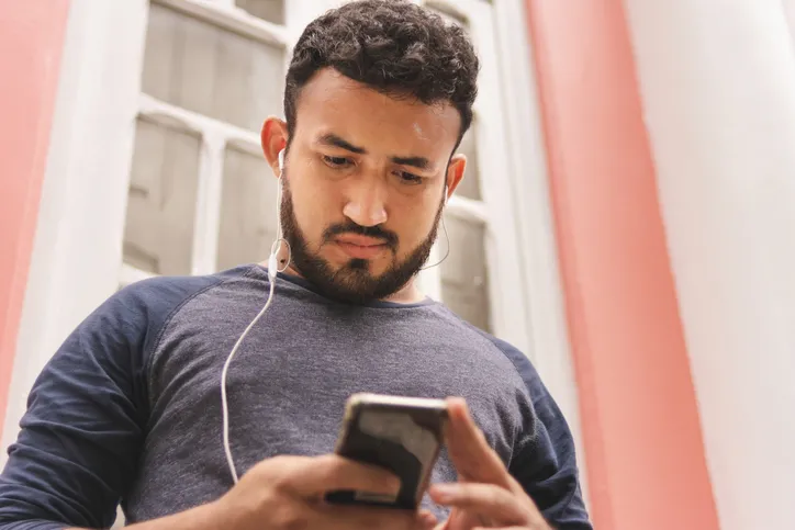 A young adult man is standing outside a house with an ear bud in while looking at his phone.