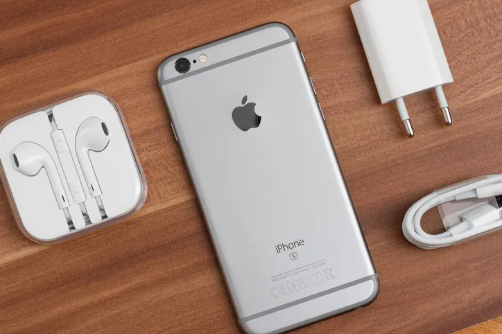 An old iPhone, Airpods and a charger sit on a brown table top.