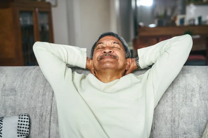 An older man is sitting on the couch with his arms behind his head while looking up and smiling.