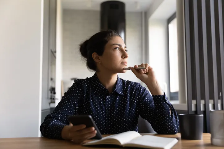 A women sitting while thinking at a kitchen table with a notebook, pen, and phone in front of her.